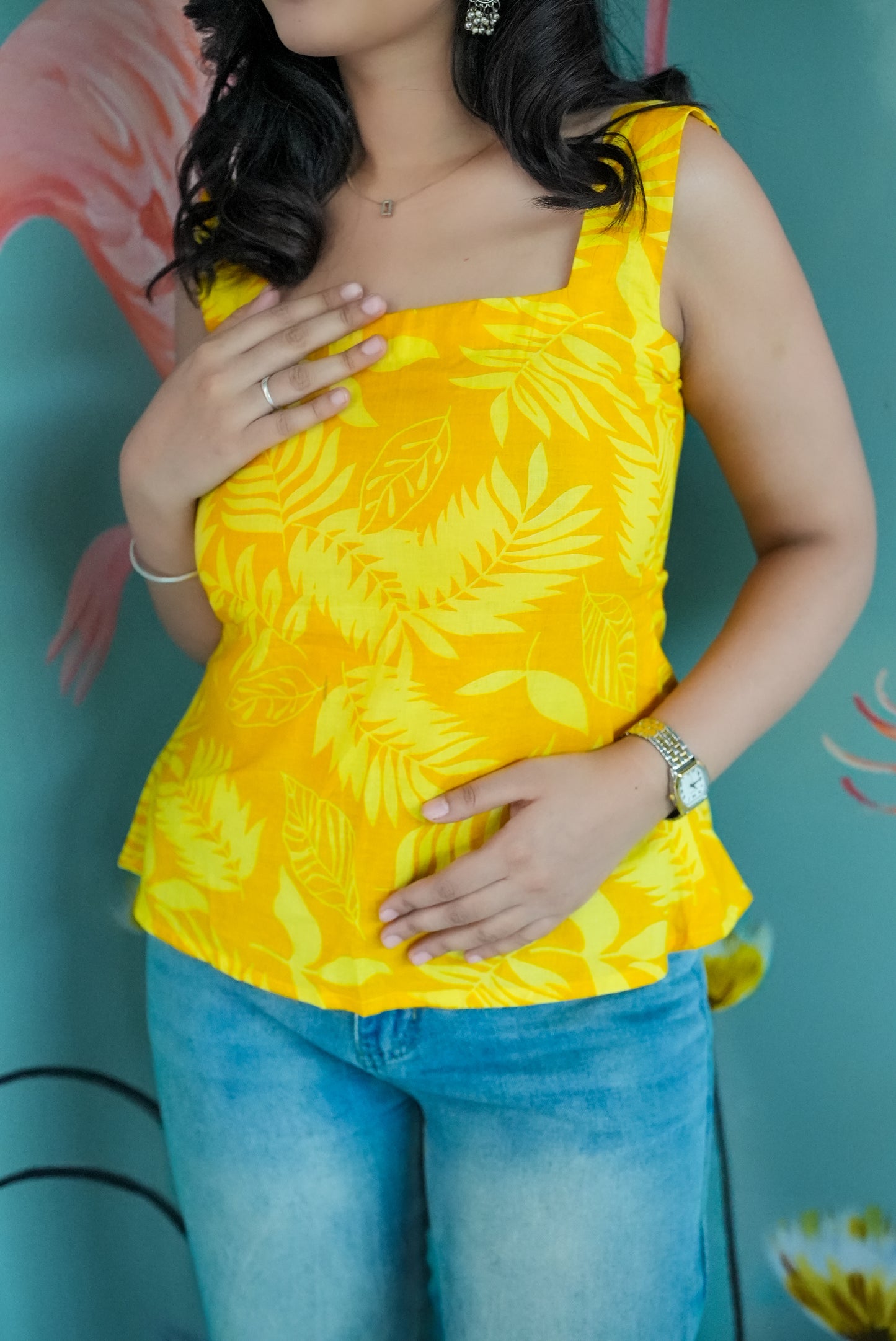 Woman wearing a yellow sleeveless top with leaf pattern and blue jeans against a teal background. Karaa