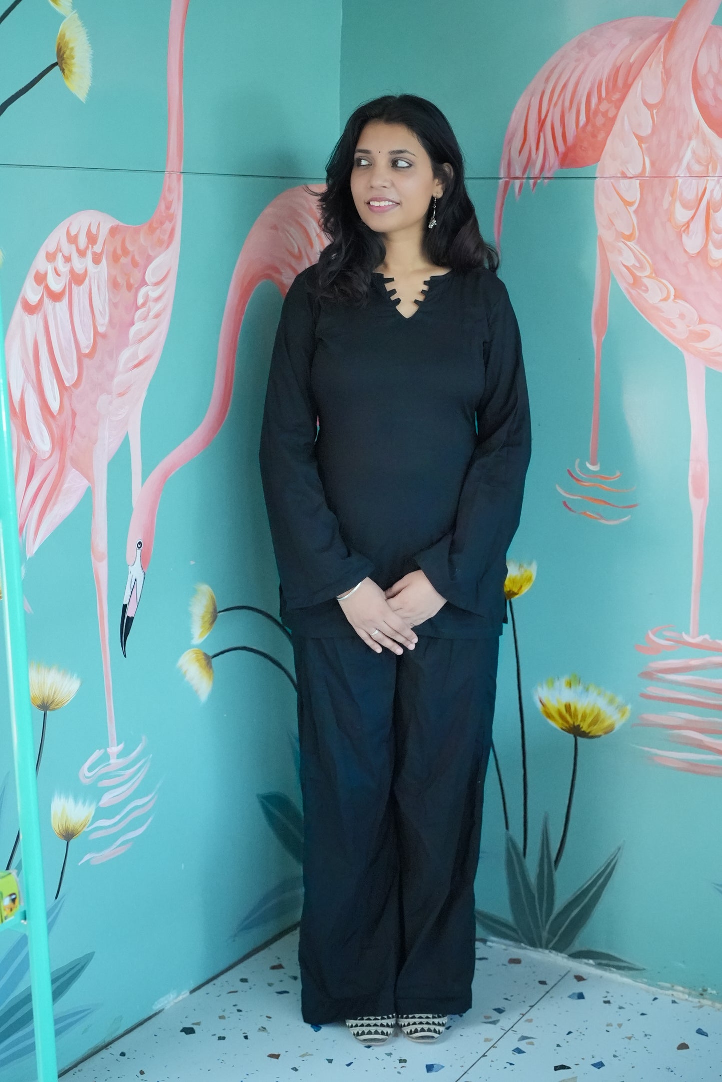 Woman in a black outfit standing in front of a colorful flamingo mural. Karaa