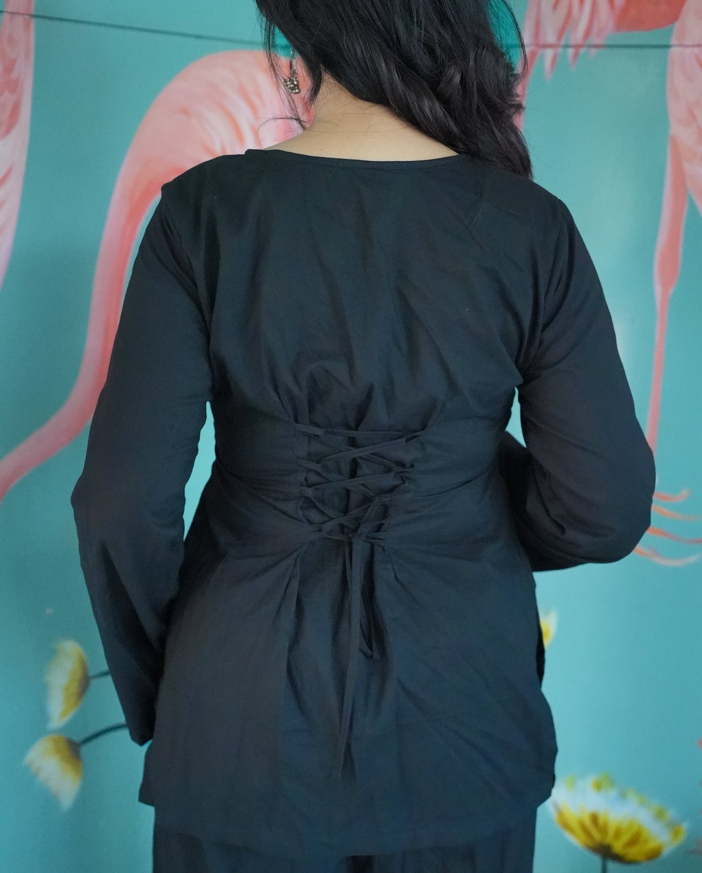 Person wearing a black outfit with a tie detail in front of a floral background 
Karaa 