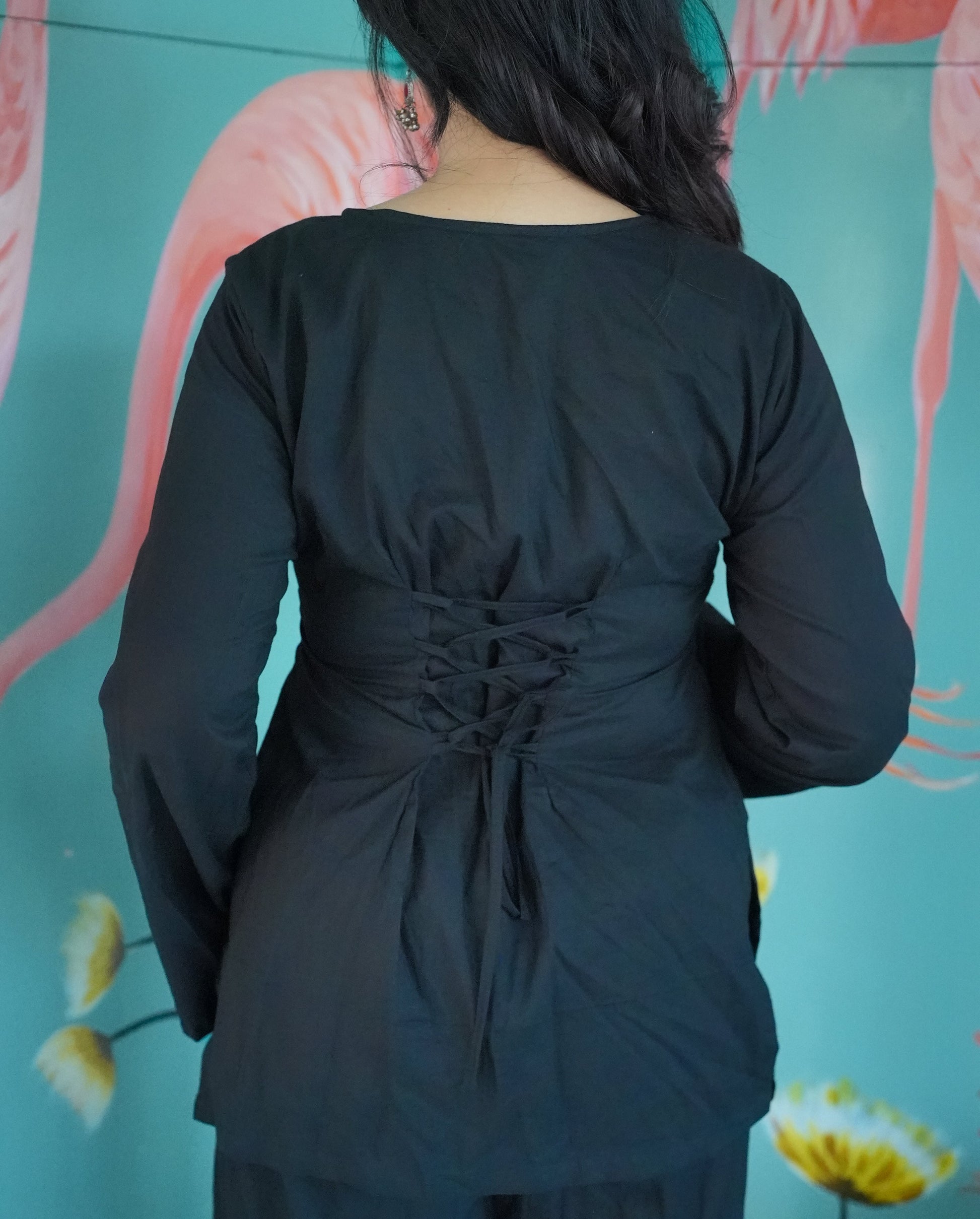Person wearing a black outfit with a tie detail in front of a floral background 
Karaa 