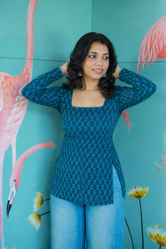 Woman in a blue patterned top and jeans standing against a teal wall with flamingo and flower illustrations. Karaa