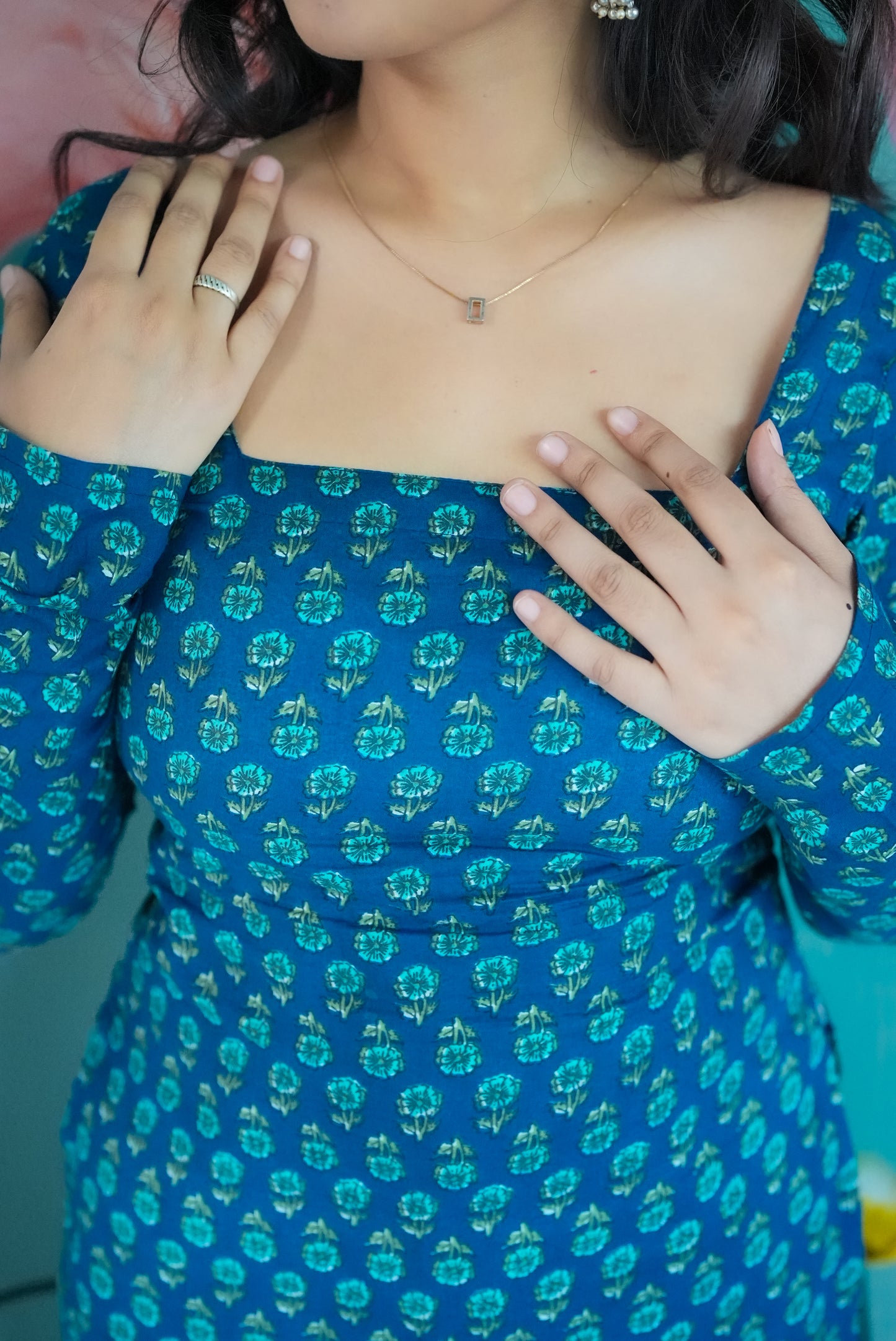Person wearing a blue dress with green floral pattern, holding chest. Karaa