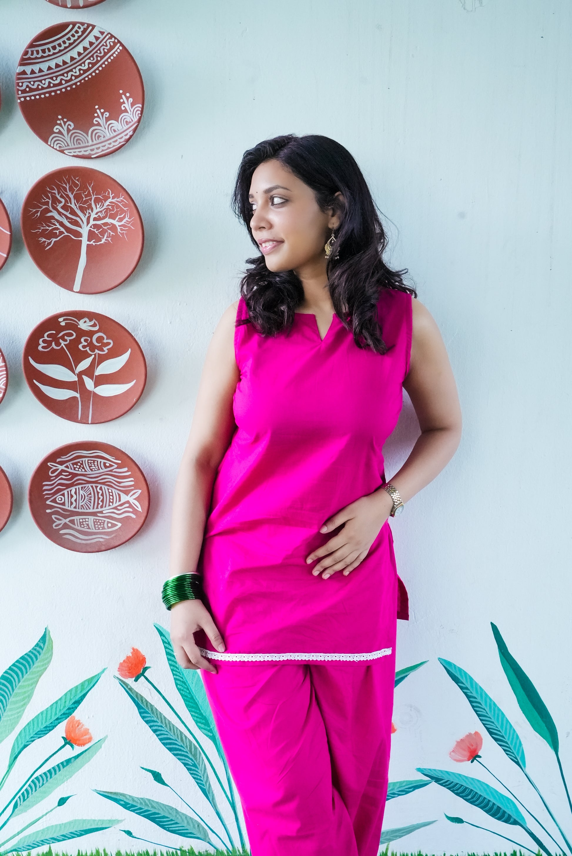 Woman in a pink outfit standing against a wall with decorative plates and floral patterns. Karaa
