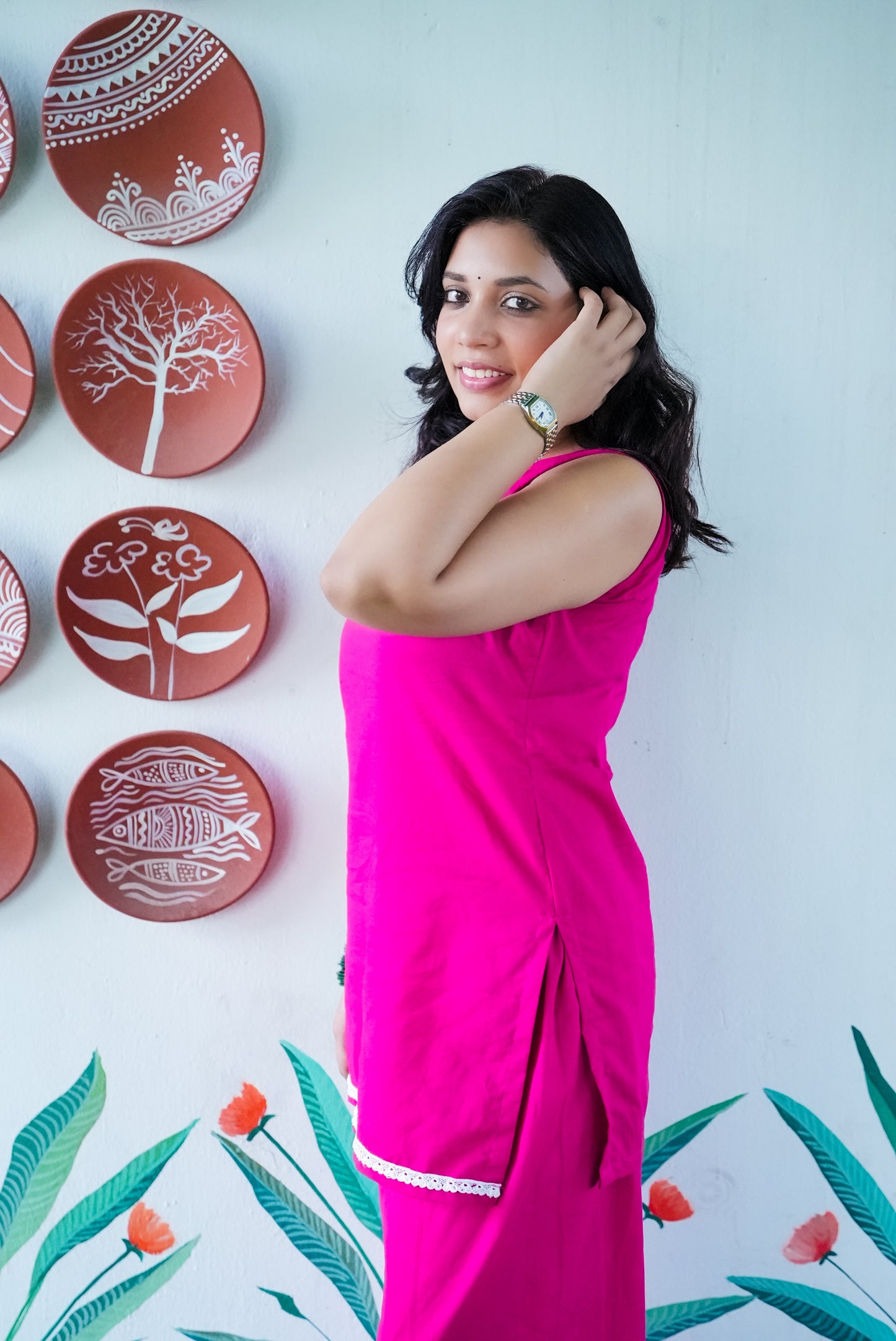 Woman in a pink outfit posing in front of decorative plates on a wall with floral designs. Karaa