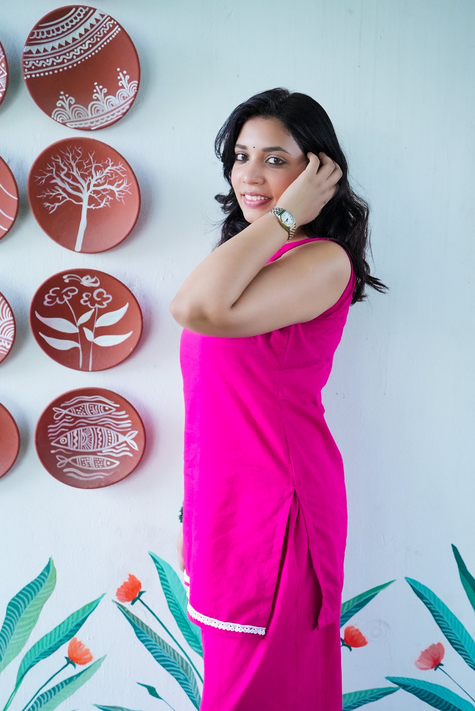 Woman in a pink outfit posing in front of decorative plates on a wall with floral designs. Karaa