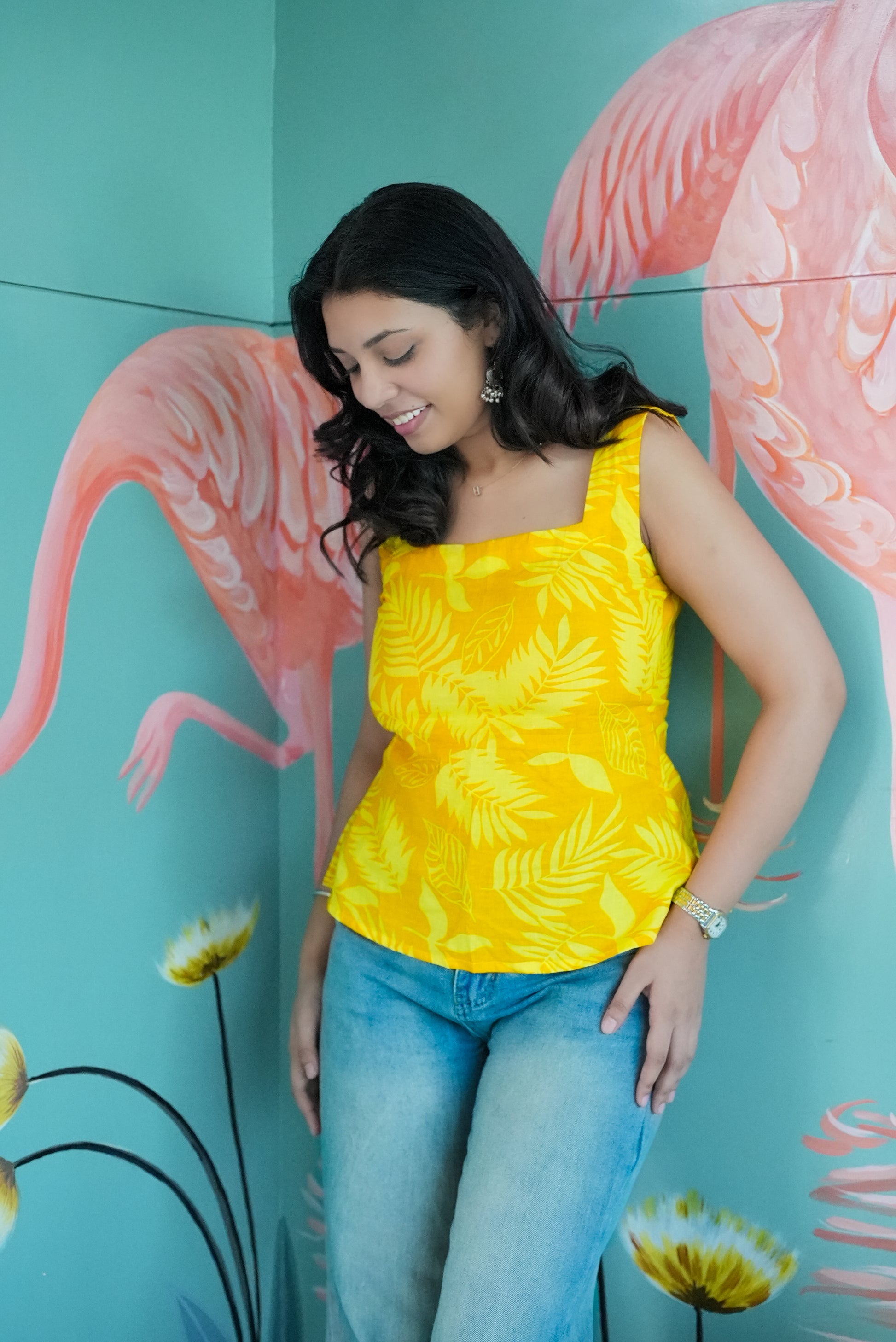 Woman wearing a yellow sleeveless top and blue jeans standing in front of a colorful flamingo mural.  Karaa