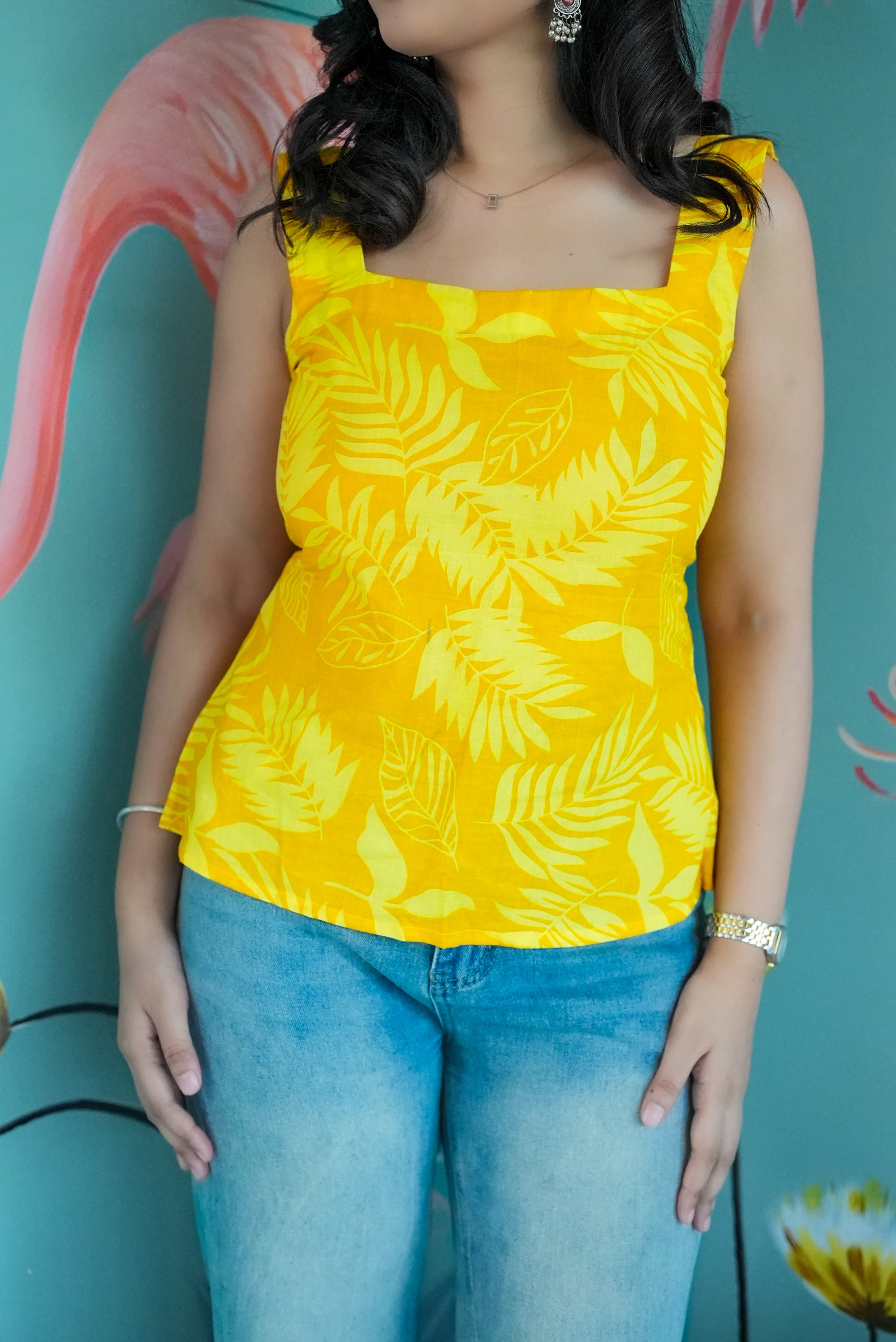 Woman wearing a yellow sleeveless top with leaf pattern and blue jeans against a teal background. Karaa