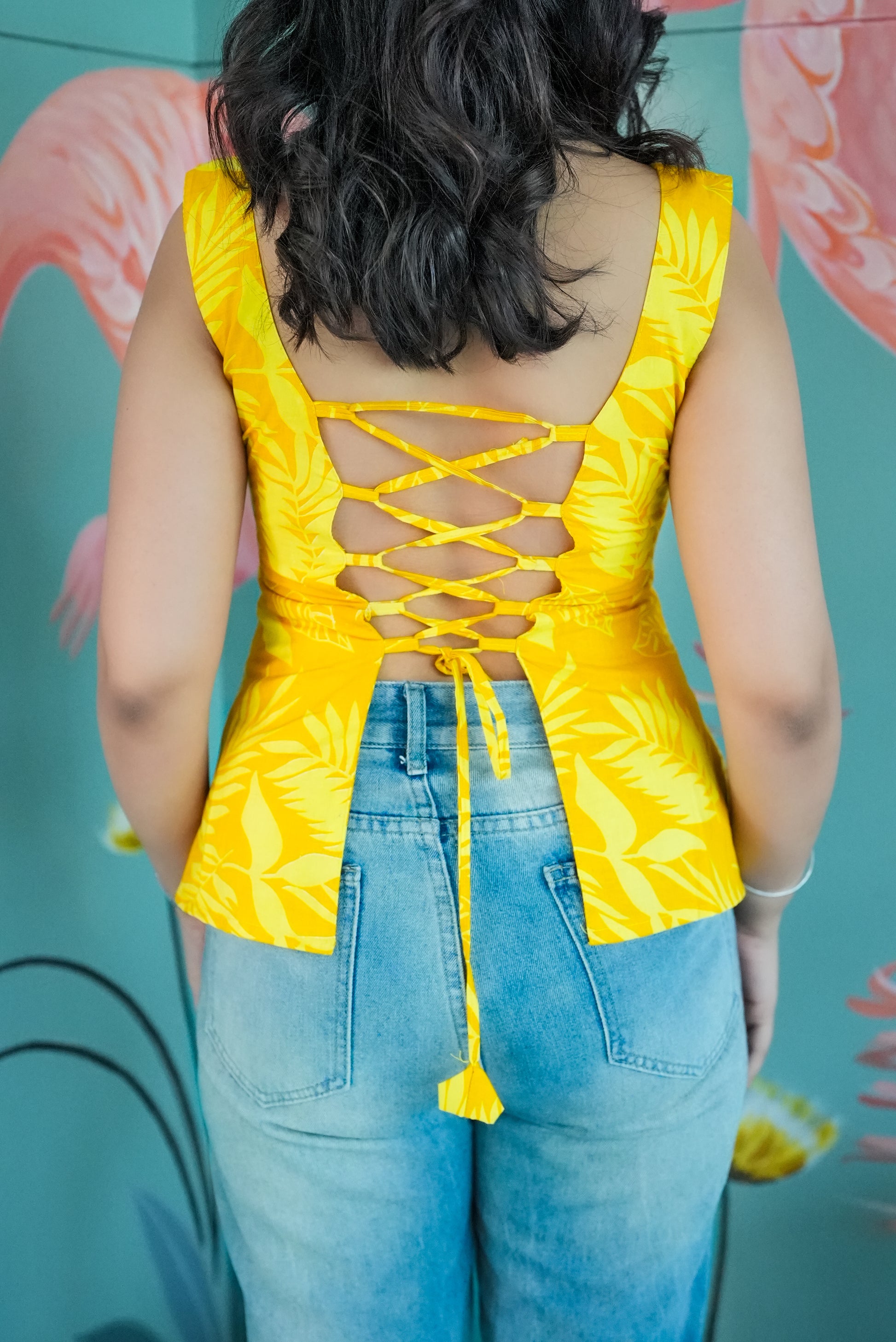 Person wearing a yellow top with a floral pattern and blue jeans, standing against a colorful background. Karaa